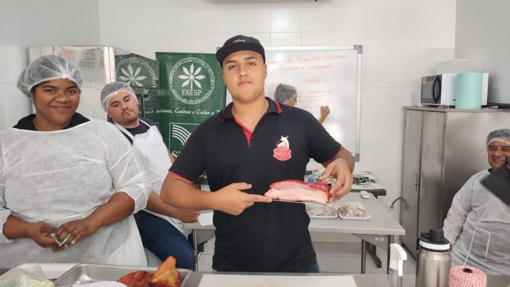 A imagem mostra um grupo de pessoas em um ambiente que parece ser uma cozinha industrial ou laboratório de alimentos, provavelmente durante um curso ou workshop sobre fabricação de produtos cárneos salgados e curados. No centro da imagem, um jovem homem está segurando uma peça grande de carne curada, exibindo-a para a câmera com um sorriso. Ele veste uma camiseta preta com detalhes vermelhos e um boné, ambos com a marca "Carnes Bardeli". Ao redor dele, há outras pessoas usando toucas e aventais brancos, indicando um ambiente higiênico e controlado, típico de manipulação de alimentos. Atrás deles, é possível ver uma parede com azulejos brancos, um quadro branco com anotações e alguns equipamentos de cozinha, como micro-ondas e geladeira. Na parte superior da imagem, há uma faixa verde com o logo da FAESP (Federação da Agricultura e Pecuária do Estado de São Paulo), sugerindo que o evento ou curso é promovido por essa entidade. Também aparecem escritos termos relacionados à fabricação de produtos cárneos, como "Jerked Beef" e "Charque", indicando que o foco do curso é o preparo de carnes salgados e curados. O ambiente transmite uma atmosfera de aprendizado prático e profissional, voltado para a produção artesanal ou industrial de carnes processadas.