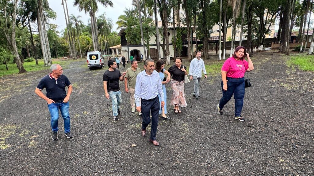 A imagem mostra um grupo de pessoas caminhando em um caminho de cascalho escuro cercado por árvores altas e folhagens exuberantes. Ao fundo, há um edifício de dois andares com uma varanda e telhado escuro, parecendo uma casa ou um hotel. Uma van branca com um símbolo vermelho e branco está estacionada mais adiante no caminho. As pessoas, vestindo roupas casuais, parecem estar em uma visita ou passeio, com algumas conversando entre si enquanto caminham. O ambiente sugere um local rural ou um parque com arquitetura distinta.