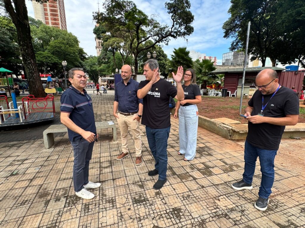A imagem mostra um grupo de cinco pessoas em um parque, em um dia nublado. No centro da imagem, um homem com camiseta preta e jeans está falando com os outros, levantando a mão direita como se estivesse gesticulando. Ele tem uma estampa na camiseta que diz "RCIÚMA Brasil". À sua esquerda, um homem com camiseta azul escura e calça jeans escura está de pé, olhando para o homem de camiseta preta. À direita do homem de camiseta preta, uma mulher com camiseta preta e calça branca está em pé, olhando para o homem de camiseta preta. Mais à direita, um homem com camiseta preta e jeans está olhando para o celular que segura nas mãos. Ele usa óculos e tem um crachá pendurado no pescoço. No lado esquerdo da imagem, um homem com camiseta azul escura e calça jeans escura está de pé, olhando para o homem de camiseta preta. Ele tem uma pedra no bolso, segurando-a com a mão direita. O chão do parque é pavimentado com pedras quadradas. Ao fundo, há árvores, um prédio alto, uma torre de igreja e um parquinho infantil.