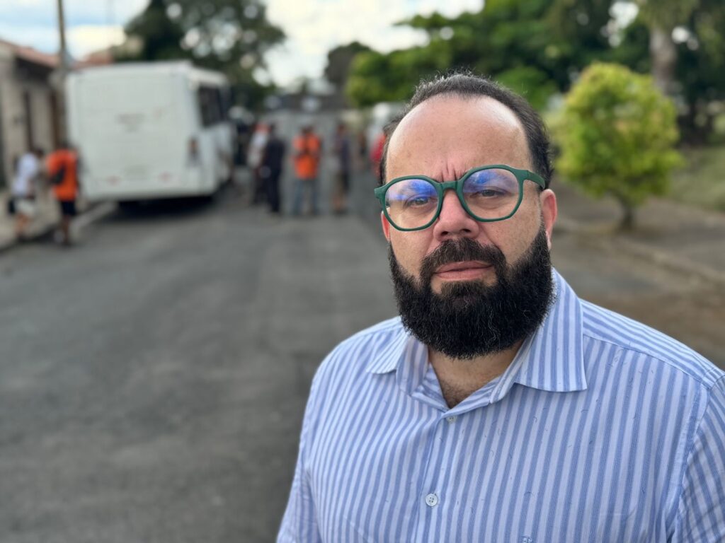 A imagem mostra um close-up de um homem com barba e óculos verdes, vestindo uma camisa listrada azul e branca. Ele está olhando diretamente para a câmera com uma expressão séria. Ao fundo, embaçado, vê-se uma rua de asfalto com algumas pessoas e um ônibus branco. As pessoas ao fundo parecem estar vestindo coletes laranja, sugerindo que podem ser trabalhadores ou voluntários em alguma atividade. Há árvores e vegetação ao redor, indicando um ambiente externo. A iluminação sugere que seja dia, com céu nublado.