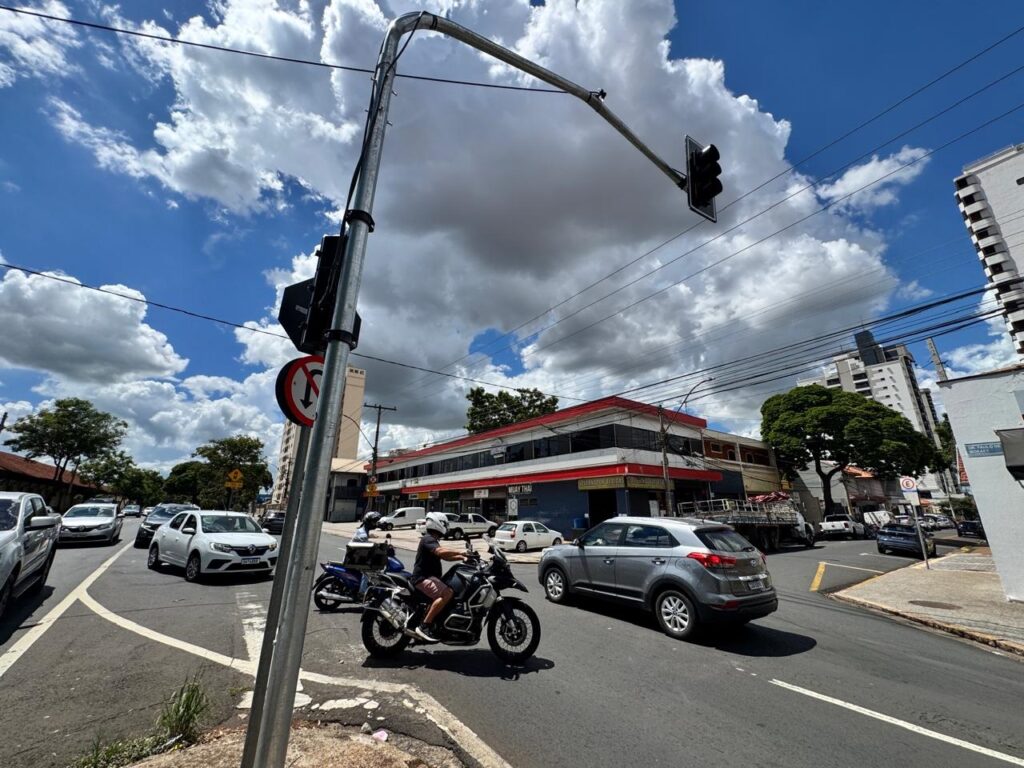 A imagem mostra uma cena de trânsito em um dia ensolarado com nuvens no céu. Vários carros e uma motocicleta estão em uma interseção com um semáforo. Ao fundo, há prédios de diferentes alturas e árvores. Um dos prédios tem uma fachada com detalhes em vermelho e branco, e há uma placa com a inscrição "GUAY THAI". Um dos carros é um SUV cinza, e a motocicleta é preta com detalhes em branco e azul. Há também placas de trânsito, incluindo uma que indica "proibido virar à esquerda". A perspectiva da câmera está baixa, dando ênfase ao semáforo e à estrutura metálica que o sustenta.