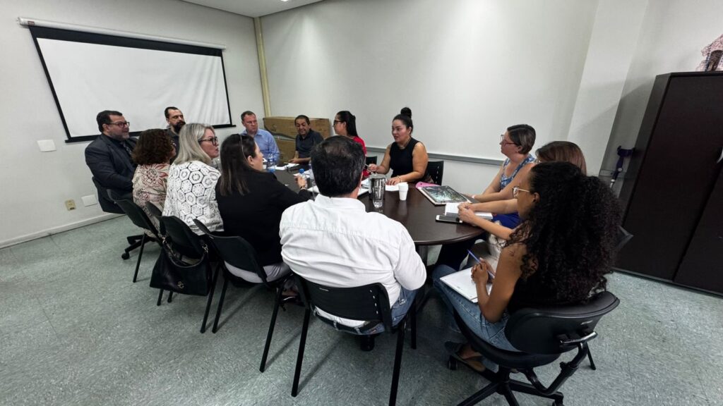 A imagem mostra uma reunião em uma sala de conferências. Um grupo de pessoas está sentado ao redor de uma mesa redonda. Há um projetor na parede atrás deles. Na mesa, há cadernos, canetas, copos e alguns documentos. As pessoas parecem estar engajadas na discussão, com algumas olhando para a frente e outras conversando entre si. A iluminação da sala é clara e uniforme, sugerindo um ambiente de escritório. O ambiente geral da imagem é profissional e formal, típico de uma reunião de trabalho ou acadêmica.