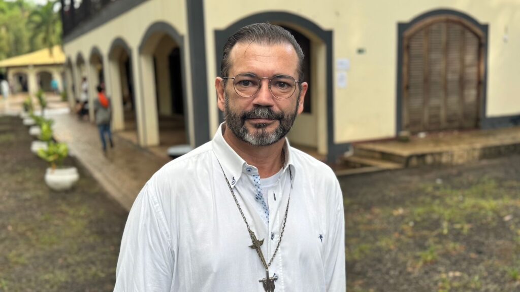 O homem na imagem está usando uma camisa branca de botões, com um medalhão em forma de cruz pendurado em uma corrente. Ele tem óculos redondos e barba, e seu cabelo está penteado para trás. Ele está em pé do lado de fora de um prédio com um telhado amarelo e colunas. Há algumas pessoas desfocadas e plantas em vasos ao redor dele. O chão está molhado, sugerindo que pode ter chovido.