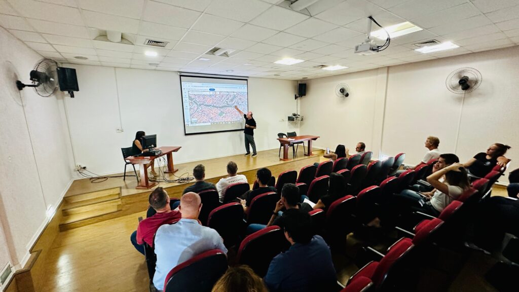A imagem mostra uma sala de aula ou auditório onde um homem está apresentando um mapa em uma tela grande. Ele está em pé na frente da tela, apontando para um ponto específico no mapa. Há uma mulher sentada em uma mesa à esquerda, olhando para um computador. Uma audiência está sentada em cadeiras vermelhas dispostas em fileiras, observando a apresentação. A sala é iluminada por luzes no teto e há ventiladores de parede. A arquitetura da sala sugere um ambiente educacional ou de conferência.