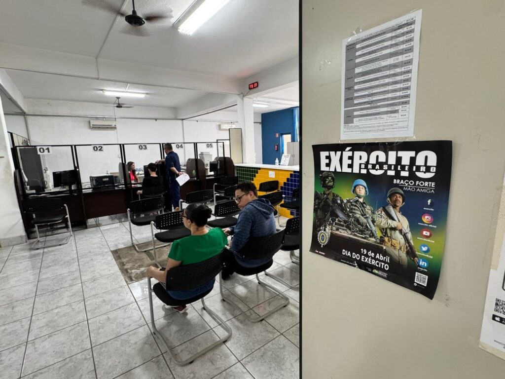 Na parte esquerda, vemos o interior de um escritório com balcões numerados de 01 a 05. Várias pessoas estão sentadas em cadeiras, aparentemente aguardando atendimento. Há computadores nos balcões e um funcionário em pé no balcão 04. O ambiente parece ser um local de serviço público. Na parte direita, um pôster grande está afixado na parede. O pôster anuncia o "EXÉRCITO BRASILEIRO" com o slogan "BRAÇO FORTE MÃO AMIGA". Abaixo, informa "19 de Abril DIA DO EXÉRCITO". O pôster também exibe ícones de redes sociais como Facebook, Instagram, YouTube e LinkedIn, indicando a presença do exército nessas plataformas. Acima do pôster, um documento com várias colunas de texto está preso à parede.