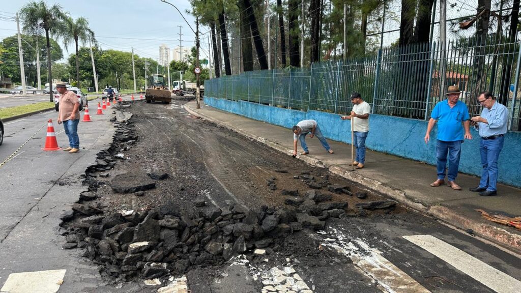 A imagem mostra uma cena de obras em uma rua durante o dia. O asfalto foi removido em uma faixa da via, revelando uma superfície de terra e pedras escuras. Há cones de sinalização laranja ao longo da área em obras para delimitar o espaço. Cinco homens estão presentes: um à esquerda próximo aos cones, outro curvado sobre o meio-fio, um terceiro com uma ferramenta na mão, e dois à direita, um deles olhando para um celular. Ao fundo, há uma máquina compactadora amarela e um caminhão branco. A rua é cercada por uma cerca azul e árvores altas, provavelmente pinheiros. Um sinal de limite de velocidade de 50 km/h está visível em um poste. O ambiente parece ser urbano ou suburbano, com iluminação natural em um dia claro, mas sem sol forte. A cena transmite uma rotina de manutenção ou reparo viário.