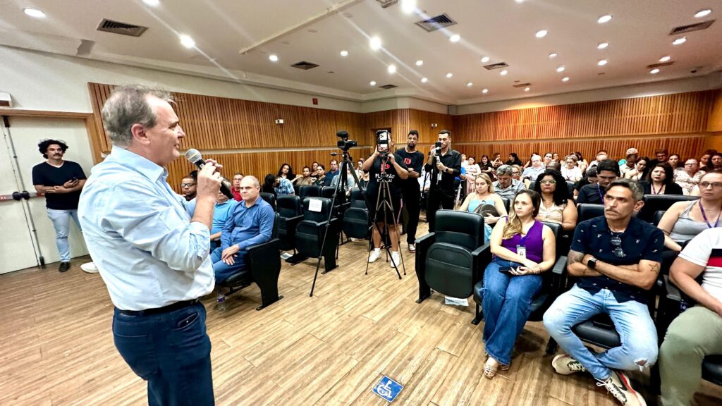 A imagem mostra uma sala de conferências com uma audiência sentada em cadeiras. Um homem no lado esquerdo da imagem está de pé, falando em um microfone e vestindo uma camisa azul clara e calças azuis. Várias pessoas na audiência estão olhando para ele, e outras estão conversando. Há também algumas pessoas em pé atrás da audiência, algumas delas segurando câmeras, o que sugere que o evento está sendo filmado ou transmitido. O ambiente parece ser formal, com paredes de madeira e iluminação de teto. Uma pequena placa azul com o símbolo de acessibilidade para cadeiras de rodas está no chão em primeiro plano.
