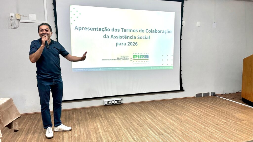 A imagem mostra um homem em pé em um chão de madeira em frente a uma tela de projeção. Ele está segurando um microfone e falando, com o braço esquerdo estendido. Ele está vestindo uma camisa polo azul escura e jeans azuis escuros, com tênis brancos. A tela de projeção exibe texto em português, que parece ser o título de uma apresentação: "Apresentação dos Termos de Colaboração da Assistência Social para 2026". Abaixo deste título, há logotipos de "SECRETARIA DE ASSISTÊNCIA, DESENVOLVIMENTO SOCIAL E FAMÍLIA" e "PREFEITURA PIRA PIRACICABA". A parte inferior da tela também contém texto menor e identificadores de redes sociais. À esquerda do homem, há uma mesa coberta com um pano. À direita, um pódio de madeira é parcialmente visível. A sala tem uma parede de cor clara com tomadas elétricas e algumas luminárias visíveis no chão.