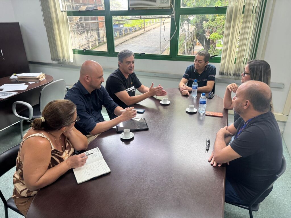 A imagem mostra um grupo de seis pessoas reunidas em torno de uma mesa de madeira em uma sala de reuniões. Há quatro homens e duas mulheres. Todos parecem estar engajados em uma discussão, com alguns gesticulando e outros ouvindo atentamente. Na mesa, há xícaras de café, garrafas de água, um tablet e um caderno com anotações. Ao fundo, uma janela com persianas revela um exterior com prédios e árvores. A sala parece ser um escritório, com arquivos e armários visíveis em um canto. A iluminação é natural, vinda da janela, e artificial, de uma lâmpada no teto. As pessoas estão vestidas de forma casual, a maioria com camisetas. Uma das mulheres está escrevendo em seu caderno, enquanto um dos homens aponta para o tablet. A atmosfera geral sugere uma reunião de trabalho produtiva.