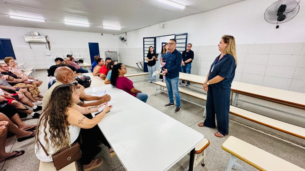 A imagem retrata uma sala com várias pessoas sentadas em mesas compridas, como se estivessem em uma reunião ou aula. No centro da sala, um homem de camisa azul e calça jeans está de pé, segurando um livro ou folder e falando com o grupo. Ao lado dele, uma mulher de uniforme escuro, possivelmente uma profissional de saúde ou educadora, também está de pé, observando a audiência. Outras pessoas estão em pé ao fundo, incluindo algumas mulheres e um homem. A sala parece ser um espaço comunitário ou educacional, com paredes brancas e azulejos, iluminação fluorescente e um ventilador de teto. As pessoas sentadas na frente parecem estar atentas à apresentação, com algumas delas segurando papéis. A atmosfera geral sugere um ambiente de aprendizado ou discussão.