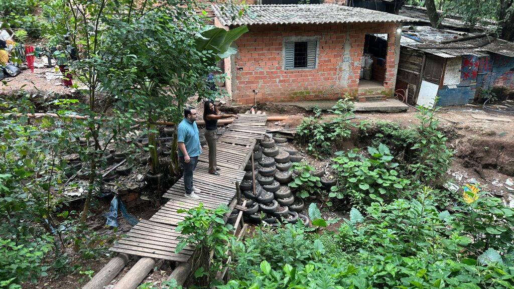 A imagem mostra um cenário que parece ser uma comunidade carente, com casas simples construídas com materiais variados, como tijolos aparentes e telhas de zinco. Há uma grande quantidade de vegetação exuberante, o que sugere um ambiente úmido e possivelmente uma área de mata ou encosta. No centro da imagem, um casal está em uma ponte de madeira improvisada, que atravessa um pequeno desfiladeiro ou vala. A ponte é construída com tábuas de madeira e apoiada em troncos na base. O chão ao redor da ponte está coberto por uma pilha de pneus usados, que parecem ter sido empilhados para conter a terra ou para algum tipo de construção informal. Ao fundo, vislumbra-se uma casa de tijolos com uma janela gradeada e, ao lado, outras construções mais precárias, feitas de madeira e metal, com telhados desgastados. Há também sinais de desordem e acúmulo de materiais diversos, como sacos plásticos e objetos. A vegetação é densa e cresce em meio aos detritos e às estruturas das casas, indicando uma convivência próxima entre a natureza e o ambiente construído.