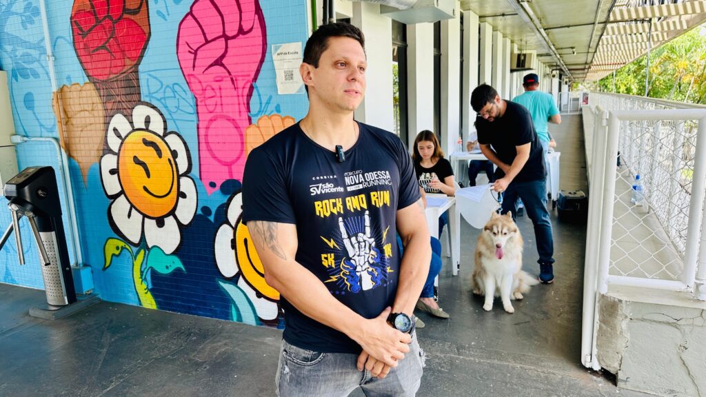 A imagem mostra um homem em primeiro plano, vestindo uma camiseta preta com uma estampa gráfica vibrante e jeans. Ele está em pé em frente a um mural colorido com desenhos de punhos cerrados e flores sorridentes. O homem está olhando para o lado, com uma expressão pensativa. Ao fundo, um corredor longo com colunas brancas e um teto coberto. Pessoas estão em mesas, possivelmente em um evento ou registro. Um homem com óculos está segurando a coleira de um cachorro husky siberiano, que está sentado no chão com a língua para fora. Mais adiante, outras pessoas e um cachorro menor são visíveis. A cena sugere um evento ao ar livre, talvez uma corrida ou feira de adoção de animais.