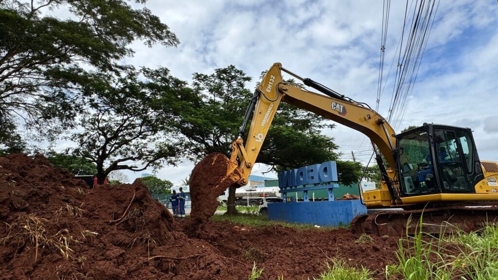 A imagem mostra um escavadeira amarela da marca CAT em operação, removendo terra vermelha em um dia nublado. A máquina está com sua concha cheia de terra, prestes a depositá-la em uma pilha recém-formada. Ao fundo, há uma placa azul com letras grandes e azuis que parecem formar uma palavra, possivelmente um nome ou marca. Ao lado da placa, há algumas árvores frondosas com folhagem verde intensa e, mais ao fundo, é possível ver fios elétricos suspensos e um caminhão tanque. Dois trabalhadores, vestidos com uniformes azuis, observam a operação da escavadeira, um deles dentro da cabine do operador. A cena sugere um trabalho de construção ou terraplanagem em andamento.