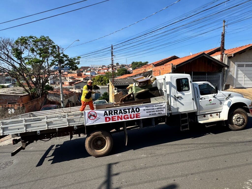 A imagem mostra um caminhão branco de coleta de lixo em uma rua suburbana, carregado com móveis velhos e outros itens. Um trabalhador vestindo um colete amarelo e calças laranjas está em pé na parte de trás do caminhão, segurando uma vara. Uma faixa branca está presa ao lado do caminhão com o texto "ARRASTÃO DENGUE" em letras grandes e vermelhas, juntamente com informações sobre a Prefeitura Municipal de Piracicaba e a Secretaria Municipal da Saúde. O caminhão também tem o logotipo "ecoTERRA". Ao fundo, há casas com telhados vermelhos e uma paisagem urbana com muitas antenas e fios. O céu está azul e claro.