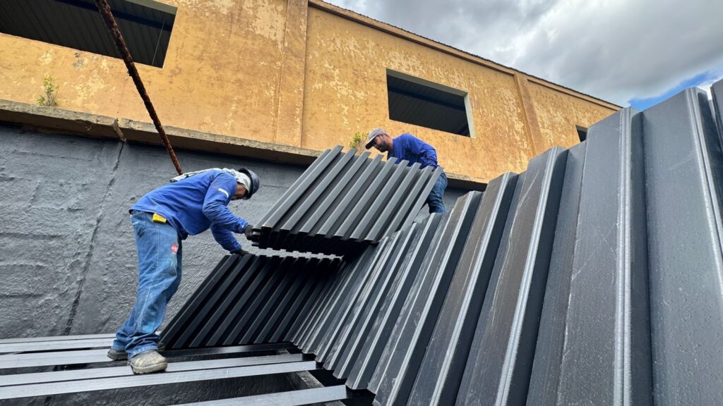 A imagem mostra dois trabalhadores em um canteiro de obras, vestindo roupas de trabalho azuis e calças jeans. Um deles está ajoelhado em primeiro plano, manipulando peças metálicas escuras e longas com ranhuras, enquanto o outro, em pé, está logo atrás, auxiliando na organização ou fixação dessas peças. As peças parecem ser parte de uma estrutura maior, possivelmente um telhado ou painel, e estão dispostas em um padrão inclinado. Ao fundo, observa-se um edifício com uma fachada desgastada em tom ocre, apresentando duas janelas retangulares e sinais de envelhecimento na pintura. Uma estrutura de concreto escuro, com uma textura áspera, estende-se pela parte inferior da imagem, servindo de base para as peças metálicas. Um cabo grosso e enferrujado, possivelmente parte de um guindaste ou equipamento de içamento, cruza a parte superior da cena, pendendo de cima. O céu, parcialmente nublado, completa o cenário, sugerindo um dia de trabalho em condições externas.
