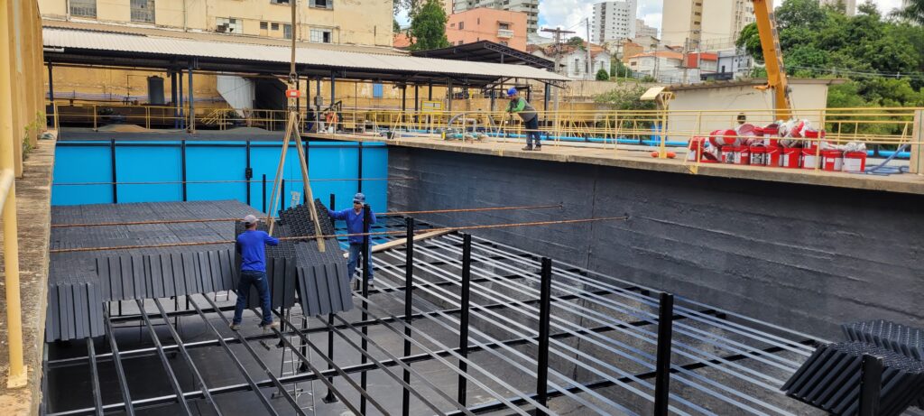 A imagem mostra uma grande estrutura de concreto, possivelmente um tanque ou reservatório, em processo de instalação de um sistema de preenchimento. Dois trabalhadores, vestidos com uniformes azuis e capacetes, estão posicionados dentro da estrutura, manipulando grandes blocos pretos que parecem ser módulos de preenchimento. Um guindaste está sendo usado para baixar mais desses blocos para a área de trabalho. Na borda superior da estrutura, há uma plataforma com corrimãos amarelos. Nessa plataforma, há vários trabalhadores supervisionando o processo e uma pilha de recipientes vermelhos, possivelmente contendo materiais de construção ou manutenção. Ao fundo, vê-se a paisagem urbana com edifícios de diferentes alturas e vegetação. As paredes internas da estrutura estão pintadas de cinza escuro, contrastando com o azul vibrante do fundo do tanque. O chão da estrutura é uma grade metálica preta.