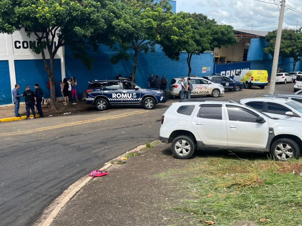 A imagem mostra uma cena em frente a um prédio com uma parede azul e a palavra "CONC" escrita em branco. Há várias pessoas em pé e alguns carros, incluindo viaturas policiais. Uma viatura azul com "ROMU" escrito na lateral está estacionada na rua. Outra viatura branca com "POLÍCIA" escrito na lateral também é visível. Há também um carro branco na frente, estacionado na grama. O céu está nublado. A cena parece ser de uma operação policial ou de segurança.