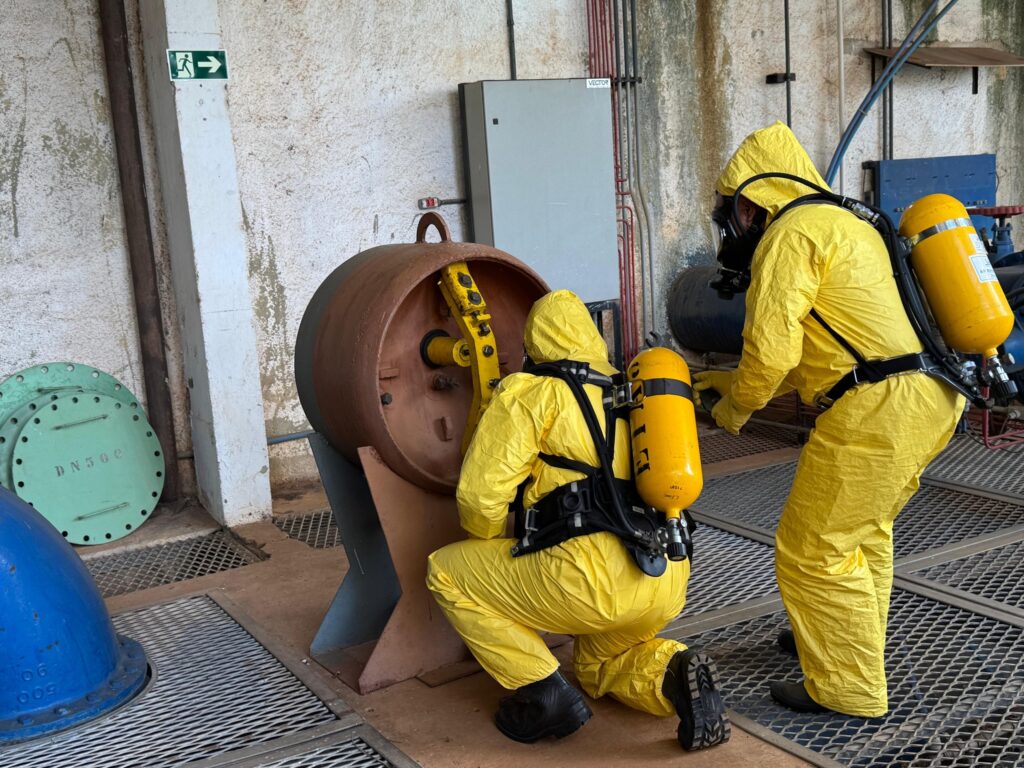 A imagem mostra duas pessoas vestindo trajes de proteção química amarelos e respiradores autônomos enquanto examinam um grande objeto cilíndrico marrom, que parece ser um equipamento industrial. Uma pessoa está ajoelhada em frente ao objeto, enquanto a outra está em pé atrás dela, ambas focadas na tarefa. O ambiente parece ser uma instalação industrial com paredes de concreto, tubulações e tampas redondas verdes no chão. Há também um sinal de saída de emergência verde com uma seta apontando para a direita na parede.