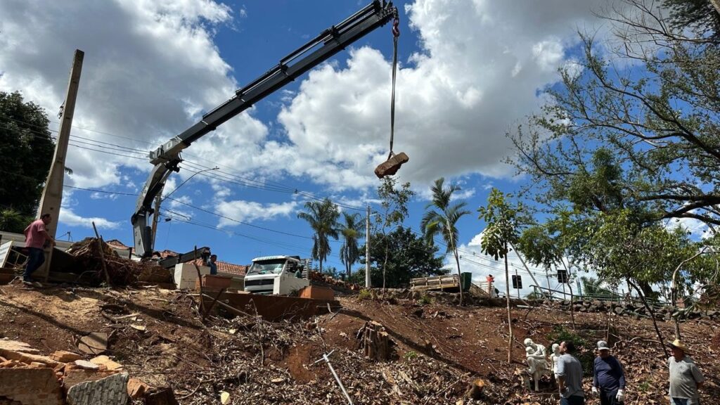 A grua está a levantar um pedaço de madeira enquanto os trabalhadores observam. O terreno está coberto de terra e destroços, sugerindo que a área foi recentemente limpa ou está em processo de reconstrução. Há uma variedade de árvores e palmeiras ao fundo, com um céu azul claro e nuvens brancas. Um camião está estacionado no lado esquerdo da imagem, possivelmente a ser carregado com os destroços. Várias pessoas, vestidas com roupas casuais, estão a observar a operação.