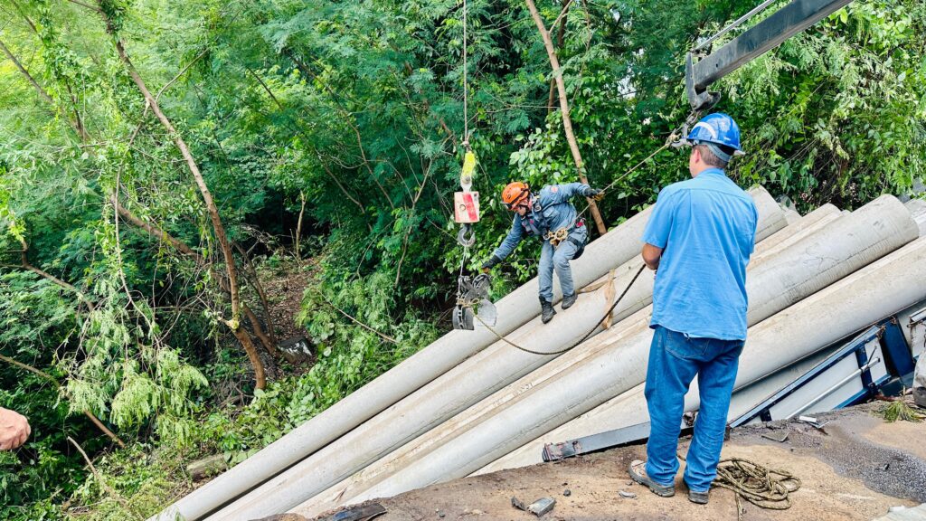 A imagem retrata um grave acidente em uma ponte, onde um caminhão tombou, espalhando sua carga de postes de concreto. A cena é de resgate e remoção, com equipes de emergência em ação.