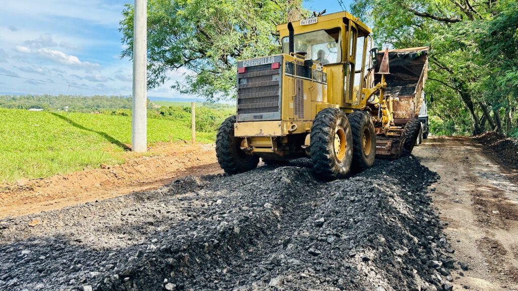 A imagem mostra um *motoniveladora Caterpillar amarela* em uma estrada de terra, espalhando material escuro, possivelmente asfalto ou um composto similar. A máquina está em primeiro plano, com suas grandes rodas sobre o material espalhado. Ao fundo, à esquerda, há um campo verde exuberante e um poste de concreto. À direita, a estrada segue por uma área arborizada, com árvores densas cobrindo o céu. O dia está ensolarado, com algumas nuvens no céu azul. A cena sugere um trabalho de manutenção ou construção de estrada em uma área rural.