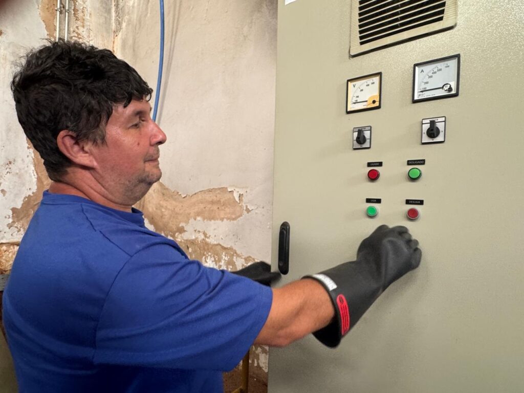 A imagem mostra um homem de perfil, vestindo uma camiseta azul e luvas pretas de proteção, interagindo com um painel de controle elétrico. O painel é de cor bege e possui diversos componentes, incluindo dois medidores (um voltímetro e um amperímetro), dois interruptores rotativos e quatro botões de comando com legendas em português: "LIGADO" (vermelho), "DESLIGADO" (verde), "LIGA" (verde) e "DESLIGA" (vermelho). O homem está com a mão direita, protegida pela luva, próxima a um dos botões. Ao fundo, observa-se uma parede com reboco descascado e um cano azul.