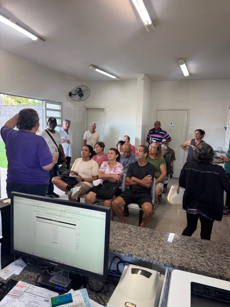 A imagem mostra uma sala de espera em uma unidade de saúde pública no Brasil, provavelmente uma UBS (Unidade Básica de Saúde). No primeiro plano, há um balcão de recepção com um computador ligado, que exibe um sistema de gerenciamento de pacientes. Sobre o balcão, há documentos e um teclado. Uma mulher vestindo uma camiseta roxa está de costas para a câmera, gesticulando em direção a um grupo de pessoas. O grupo, composto por cerca de 10 a 12 pessoas, em sua maioria adultos e idosos, está sentado em cadeiras ou em pé, prestando atenção na mulher de roxo ou em algum ponto próximo a ela. A sala possui paredes brancas, iluminação fluorescente no teto e um ventilador fixado na parede. Ao fundo, há duas portas, uma com a placa "BANHEIRO MASCULINO" e outra com a placa "SANITÁRIO PCD" (para pessoas com deficiência). À direita, uma mulher com jaqueta preta e bolsa branca caminha para fora do quadro. O ambiente transmite uma sensação de rotina típica em uma clínica de saúde pública, com pessoas aguardando atendimento ou informações. A iluminação é clara e uniforme, e a foto foi tirada de um ângulo amplo, atrás do balcão da recepção.