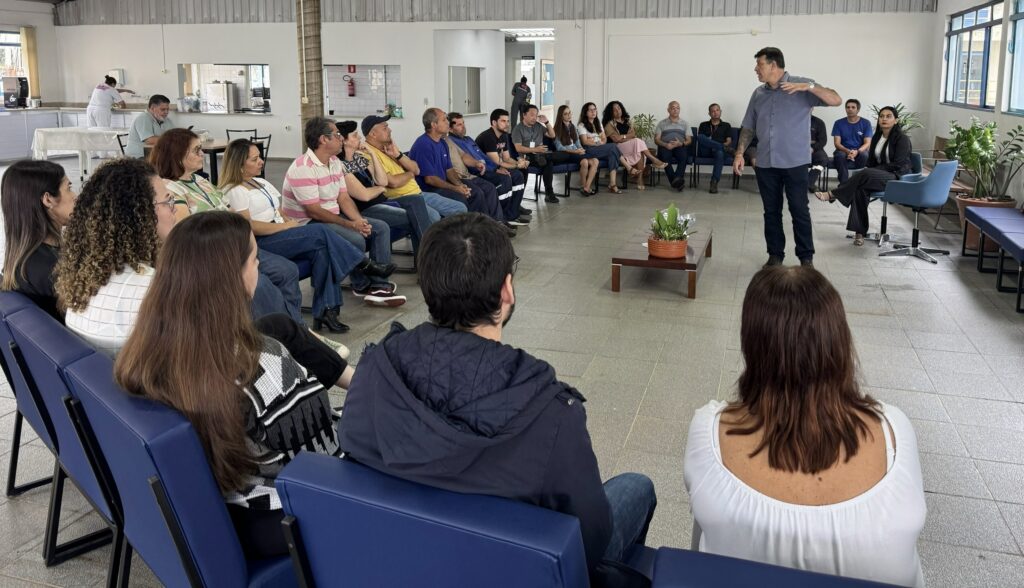 A imagem mostra um grupo de cerca de 25 a 30 pessoas reunidas em um ambiente interno amplo, provavelmente para uma reunião ou apresentação. Um homem, que parece ser o palestrante, está em pé à direita da imagem, usando uma camisa cinza de mangas curtas e calça escura, gesticulando enquanto fala para o público. As pessoas estão sentadas em cadeiras azuis e bancos, formando um semicírculo ao redor de uma pequena mesa de centro de madeira com uma planta em cima. O público é diversificado, com homens e mulheres de várias idades, vestidos de forma casual e semi-casual. O espaço tem um aspecto funcional, com piso de cerâmica cinza, paredes brancas e um teto alto de metal ondulado. Ao fundo, há uma área que parece ser uma cozinha ou cafeteria, e do lado direito, grandes janelas permitem a entrada de luz natural, iluminando bem o ambiente. A imagem transmite a sensação de um encontro informal, porém organizado, com foco na interação entre o palestrante e os participantes.
