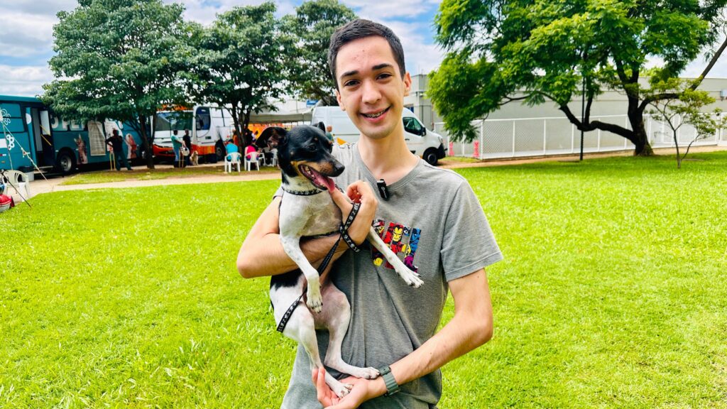 A imagem mostra um jovem sorrindo e segurando um pequeno cachorro preto e branco no colo. Ambos estão em um gramado verde com árvores ao fundo. O jovem usa uma camiseta cinza com estampas coloridas de super-heróis. O cachorro parece feliz, com a língua para fora. Ao fundo, há ônibus, pessoas e uma cerca branca. O céu está nublado.