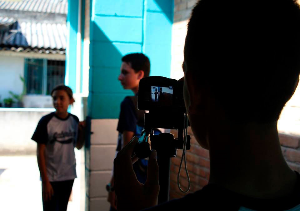 A imagem mostra uma cena de bastidores de uma gravação, onde uma pessoa está operando uma câmera digital montada em um tripé. A pessoa que manuseia a câmera aparece em silhueta, com forte contraste de luz e sombra. Em segundo plano, um jovem está de perfil, olhando para a esquerda, enquanto uma jovem está mais ao fundo, levemente desfocada, também voltada para a câmera. O ambiente parece ser ao ar livre, possivelmente em uma escola ou pátio, com uma parede azul e branca iluminada pelo sol e um telhado de cerâmica visível ao fundo. A imagem transmite a sensação de um projeto audiovisual em andamento, talvez uma atividade escolar ou comunitária.