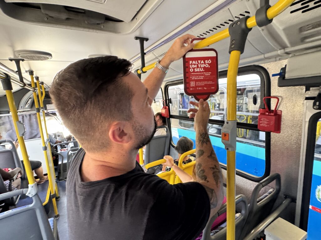 A imagem mostra o interior de um ônibus, onde um homem com barba e tatuagens no braço está pendurando um pequeno cartaz vermelho em uma barra amarela de apoio. O cartaz contém um texto em português que diz "FALTA SÓ UM TIPO, O SEU." e informações sobre o Hemocentro de Piracicaba, incentivando a doação de sangue. Há outras pessoas sentadas nos bancos do ônibus ao fundo, e as barras de apoio e postes amarelos são característicos de veículos de transporte público.