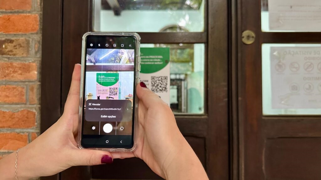 A imagem mostra uma pessoa segurando um smartphone com a câmera voltada para um QR code afixado em uma porta de vidro. O smartphone está em modo de câmera, e uma sobreposição na tela exibe informações sobre o QR code escaneado, incluindo um texto em português "Vincular" e um link para um formulário. O QR code está em um cartaz verde e branco, com outros cartazes com instruções e símbolos visíveis na porta de vidro ao fundo. A porta parece ser de madeira escura, dividida em painéis de vidro. Ao lado da porta, há uma parede de tijolos aparentes. A iluminação sugere que a cena ocorre durante o dia.