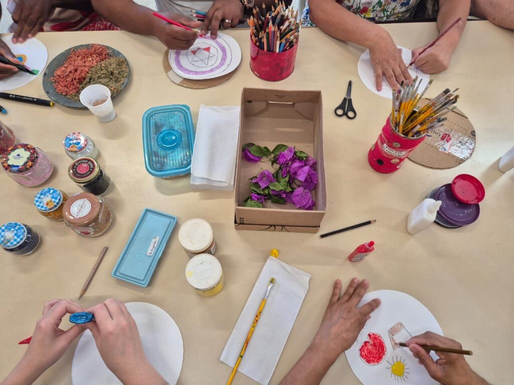 A imagem mostra uma mesa de artesanato com várias pessoas trabalhando em projetos criativos. Há potes de glitter de várias cores, tesouras, lápis de cor, pincéis e materiais para colagem, como flores roxas e grãos coloridos. As pessoas estão desenhando em papéis redondos, algumas usando lápis e outras pincéis. Uma pessoa está aplicando glitter em um projeto, enquanto outra está desenhando um sol e um coração. A cena transmite uma atmosfera de criatividade e aprendizado, com todos concentrados em suas atividades.