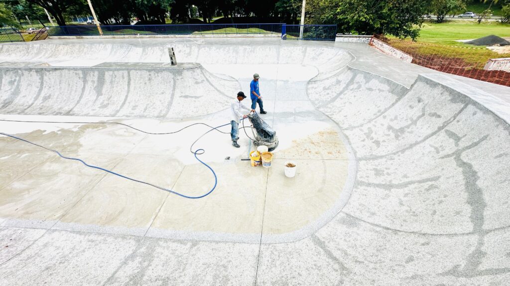 A imagem mostra uma cena aérea de uma pista de skate recém-construída, com dois trabalhadores realizando o polimento do concreto. A pista apresenta curvas amplas e superfícies inclinadas, típicas de skateparks. Um dos trabalhadores está operando uma máquina de polimento, enquanto o outro observa. Há baldes com materiais de construção próximos à máquina. A área ao redor da pista está em construção, com cercas de proteção laranja e pilhas de areia. Árvores e vegetação cercam o local, e ao fundo, uma estrada com carros pode ser vista. O concreto da pista parece ter diferentes acabamentos, com algumas áreas mais lisas e outras com uma textura mais granulada.