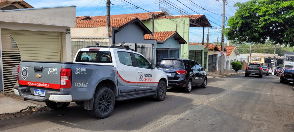 A imagem mostra uma rua residencial com várias casas e carros estacionados. Em primeiro plano, há uma picape branca e cinza com a inscrição "POLÍCIA CIENTÍFICA" e "SENASP" em suas laterais, indicando que é um veículo oficial. A picape tem o número de licença "SGQ5J28". Ao lado da picape, um carro preto está estacionado. Mais adiante na rua, outros carros e casas podem ser vistos. O céu está parcialmente nublado. Há também uma placa de trânsito indicando "20" (provavelmente o limite de velocidade) e postes de eletricidade com muitos fios. A cena parece ser de uma operação ou investigação policial em andamento.