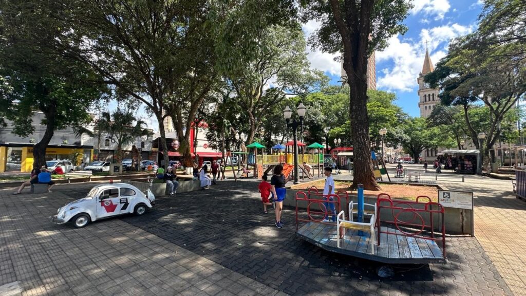 A cena retrata um dia ensolarado em uma praça pública movimentada. No primeiro plano, um pequeno carro branco, que parece ser um modelo antigo do Fusca, está estacionado. Ele possui adesivos vermelhos e pretos nas laterais e um giroflex vermelho no teto. Um carrossel infantil de madeira, com detalhes em metal vermelho, ocupa a parte inferior direita da imagem. Ao fundo, crianças e adultos circulam pela praça. Há um playground colorido com escorregadores e balanços, onde algumas crianças brincam. Árvores frondosas com copas verdes oferecem sombra em boa parte da praça, contrastando com o céu azul e algumas nuvens brancas. Ao fundo, à direita, ergue-se a torre de uma igreja com um relógio, e à esquerda, prédios comerciais e residenciais compõem o cenário urbano. Pessoas sentadas em bancos e muretas aproveitam o dia.