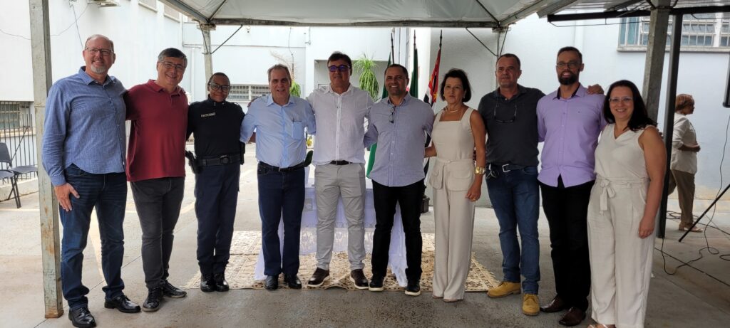 A imagem mostra um grupo de dez pessoas, sete homens e três mulheres, posando para uma foto em um ambiente externo, possivelmente um pátio ou área de eventos. Eles estão em pé, alinhados lado a lado, e parecem estar em um evento formal ou oficial, dada a presença de bandeiras ao fundo. A maioria das pessoas está vestida de forma casual a semi-formal. À esquerda, um homem com óculos e camisa azul clara está abraçando um homem com camisa vermelha. Ao lado deles, uma mulher com óculos e uniforme escuro, possivelmente de segurança ou oficial, está com o braço sobre o ombro do homem de camisa vermelha. Em seguida, há um homem com camisa azul clara e calça jeans, seguido por um homem com camisa branca e óculos. No centro do grupo, uma mulher com um conjunto de calça e blusa bege está sorrindo. Ela está cercada por homens em trajes casuais. Um homem com camisa cinza escuro e calça jeans está ao seu lado, e à sua direita, um homem com camisa lilás e calça preta. Mais à direita, uma mulher com um conjunto bege semelhante, também sorrindo, fecha o grupo principal. Ao fundo, à extrema direita, uma mulher com camisa branca está de pé, ligeiramente afastada do grupo principal. O ambiente parece ser sob uma cobertura, com postes de metal sustentando a estrutura. Ao fundo, vê-se uma parede branca com janelas e algumas plantas. As bandeiras ao fundo sugerem um contexto cívico ou institucional.