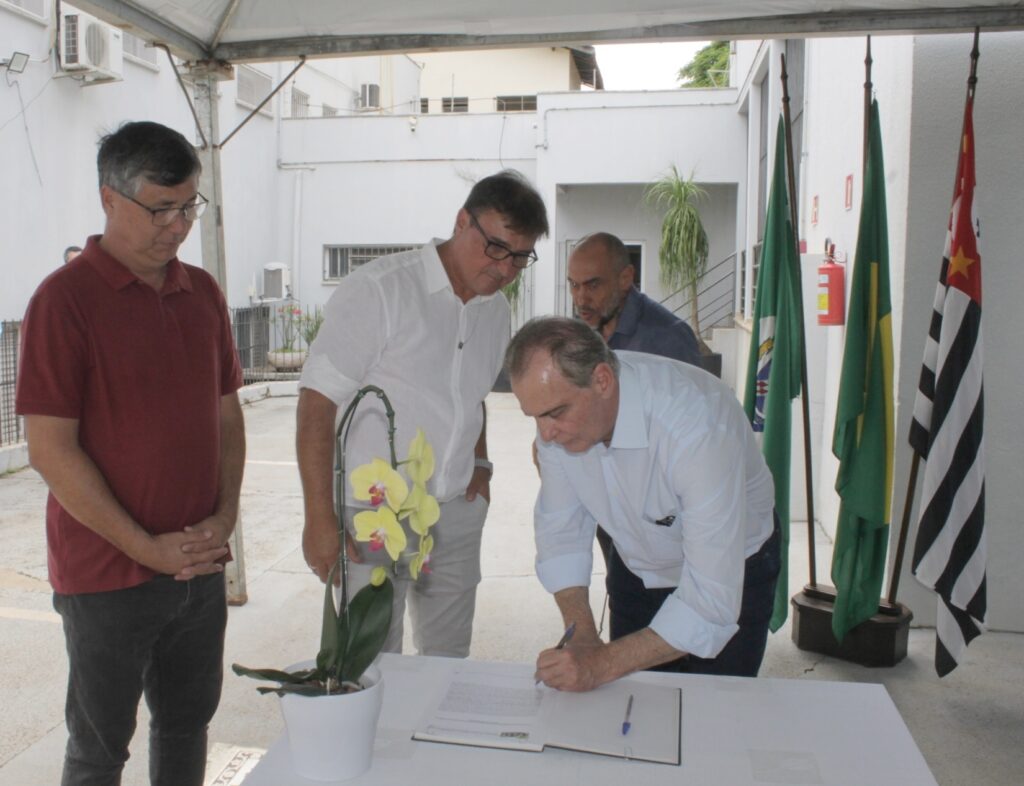 A imagem mostra um evento formal ao ar livre, possivelmente uma cerimônia de assinatura. Quatro homens estão presentes, com um deles curvado sobre uma mesa para assinar um documento. O homem que está assinando veste uma camisa social azul clara e está focado no documento à sua frente. Ao seu lado, um homem com uma camisa branca e óculos também parece observar o documento. Outro homem, vestindo uma camisa polo vinho e óculos, está de pé à esquerda, com as mãos juntas. Um quarto homem, com a cabeça raspada, está em segundo plano, atrás do homem de camisa branca. Atrás dos homens, há várias bandeiras, incluindo a bandeira do estado de São Paulo (com listras pretas e brancas e um fundo vermelho com uma estrela) e bandeiras verdes com elementos não identificáveis. Uma planta com flores amarelas em um vaso branco está colocada sobre a mesa, perto dos homens. Ao fundo, observa-se a fachada de um edifício branco com janelas e um ar condicionado. O ambiente parece ser um pátio ou uma área externa de uma instituição.