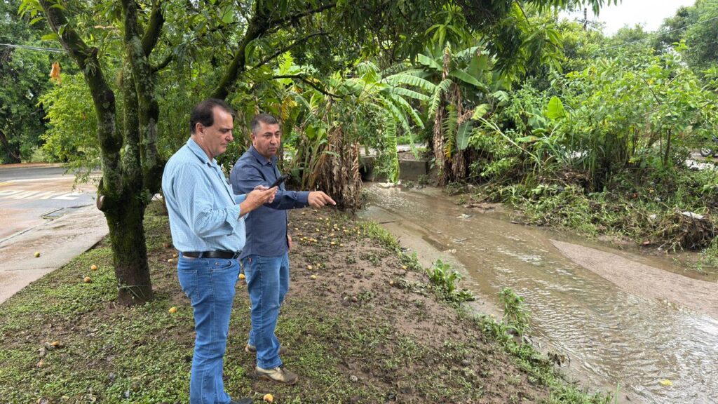A imagem mostra dois homens adultos em uma área rural, à beira de um pequeno córrego ou vala com água turva. O homem à esquerda está olhando para um celular, vestindo uma camisa social azul clara de manga longa e calça jeans. O homem à direita, que usa uma camisa azul escura e calça jeans, está apontando para algo no córrego, possivelmente para a água ou para a estrutura de concreto que aparece ao fundo, que parece ser um bueiro ou uma passagem de água sob uma estrada. O chão ao redor está lamacento e coberto por folhas e frutos amarelos caídos, indicando um ambiente úmido e talvez recente chuva. A vegetação ao redor é densa e tropical, com árvores de banana e mangueiras. Ao fundo, é possível ver uma estrada asfaltada com marcações brancas. O cenário sugere uma inspeção ou avaliação da situação do córrego ou da infraestrutura local.