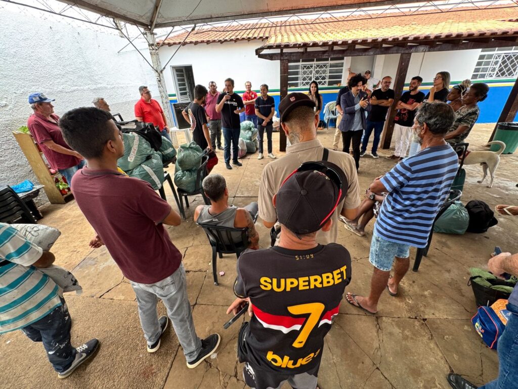 A imagem mostra um grupo de pessoas em um pátio, possivelmente em uma área externa coberta. Há várias sacolas verdes empilhadas, algumas em cadeiras. Um homem no centro, vestindo uma camiseta preta com a palavra "SUPERBET" e o número 7, está de costas para a câmera, mas sua cabeça está virada para a esquerda. Ele usa um boné preto. Outras pessoas estão em pé e sentadas ao redor. Um homem de camisa azul está falando com o grupo, segurando um celular na mão. Há uma cadela branca no lado direito da imagem, perto de algumas sacolas. O ambiente parece ser um local comunitário ou de apoio, com materiais sendo distribuídos ou organizados.