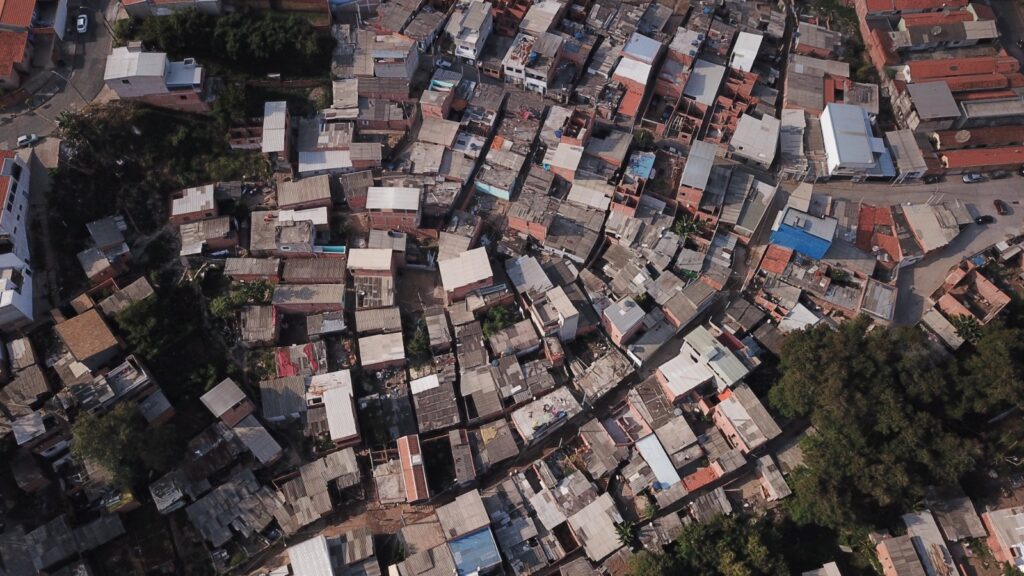 A imagem mostra um cenário que parece ser uma comunidade carente, com casas simples construídas com materiais variados, como tijolos aparentes e telhas de zinco. Há uma grande quantidade de vegetação exuberante, o que sugere um ambiente úmido e possivelmente uma área de mata ou encosta. No centro da imagem, um casal está em uma ponte de madeira improvisada, que atravessa um pequeno desfiladeiro ou vala. A ponte é construída com tábuas de madeira e apoiada em troncos na base. O chão ao redor da ponte está coberto por uma pilha de pneus usados, que parecem ter sido empilhados para conter a terra ou para algum tipo de construção informal. Ao fundo, vislumbra-se uma casa de tijolos com uma janela gradeada e, ao lado, outras construções mais precárias, feitas de madeira e metal, com telhados desgastados. Há também sinais de desordem e acúmulo de materiais diversos, como sacos plásticos e objetos. A vegetação é densa e cresce em meio aos detritos e às estruturas das casas, indicando uma convivência próxima entre a natureza e o ambiente construído.