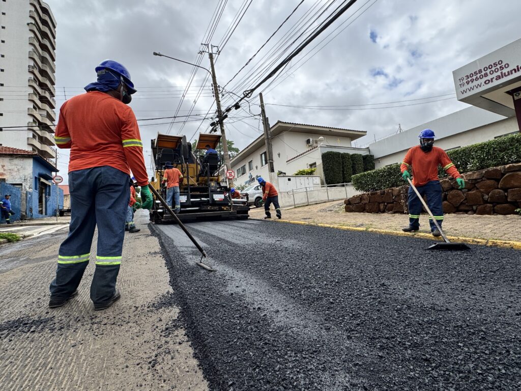 A imagem mostra uma equipe de trabalhadores realizando a pavimentação de uma rua. No primeiro plano, um trabalhador de costas, vestindo uniforme laranja de manga longa, calça jeans e capacete azul, manuseia um ancinho sobre o asfalto recém-colocado. Outros trabalhadores estão próximos a uma máquina pavimentadora amarela, que está espalhando o asfalto. A cena ocorre em uma área urbana com prédios residenciais e vegetação ao fundo, sob um céu nublado. Há também uma placa com informações de contato e um site.