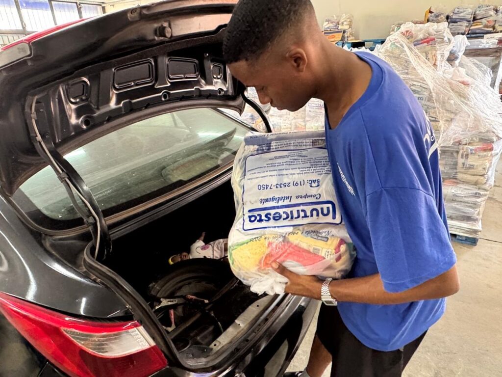 Um jovem de camiseta azul está colocando um pacote grande de alimentos em um porta-malas aberto de um carro. O pacote, que parece ser uma cesta básica, está embalado em plástico transparente e tem a marca "Nutricesto" visível. Ao fundo, há pilhas de outras cestas básicas embaladas em plástico, sugerindo que a cena se passa em um local de distribuição de alimentos, como um armazém ou centro de doações. O porta-malas do carro está escuro, e dentro dele, no canto inferior esquerdo, é possível ver uma boneca caída. O jovem usa um relógio no pulso esquerdo e parece concentrado em sua tarefa.