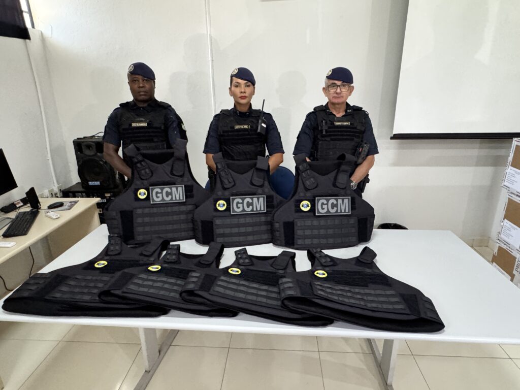 A imagem mostra três agentes da Guarda Civil Municipal (GCM) posando atrás de uma mesa onde estão expestes vários coletes balísticos táticos pretos. Os agentes estão uniformizados com roupas azul-marinho, coletes táticos e boinas da mesma cor. Eles estão alinhados, olhando diretamente para a câmera com expressões sérias e profissionais. Na mesa, há sete coletes ao todo: três estão em pé, apoiados contra a mesa, e quatro estão deitados na frente. Cada colete possui a sigla "GCM" em destaque, além de um brasão redondo com cores amarelo e azul. O ambiente parece ser uma sala interna, possivelmente um escritório ou sala de reuniões, com paredes brancas, um projetor ao fundo e um computador em uma mesa lateral. A iluminação é clara e uniforme, sugerindo luz artificial. A cena transmite a ideia de uma apresentação ou demonstração dos equipamentos de proteção utilizados pela GCM.