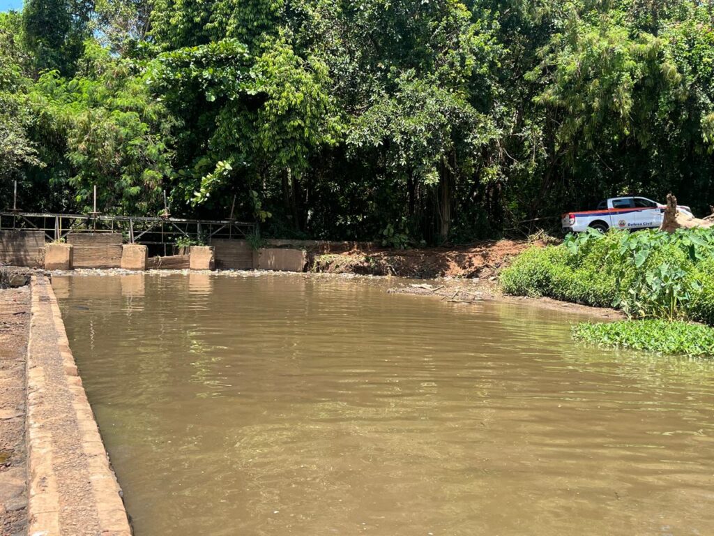 A imagem mostra uma pequena estrutura de contenção de água em meio a uma área florestada, formada por barreiras de concreto e corrimão metálico que regulam o fluxo de um rio ou canal de águas escuras e barrentas. À direita, um veículo branco da Defesa Civil está estacionado próximo à vegetação, sinalizando a presença de equipes de monitoramento ou manutenção. O cenário é cercado por densa mata verde, sugerindo um ambiente tropical ou subtropical, onde a infraestrutura de controle hídrico se integra às ações de segurança e prevenção de desastres.