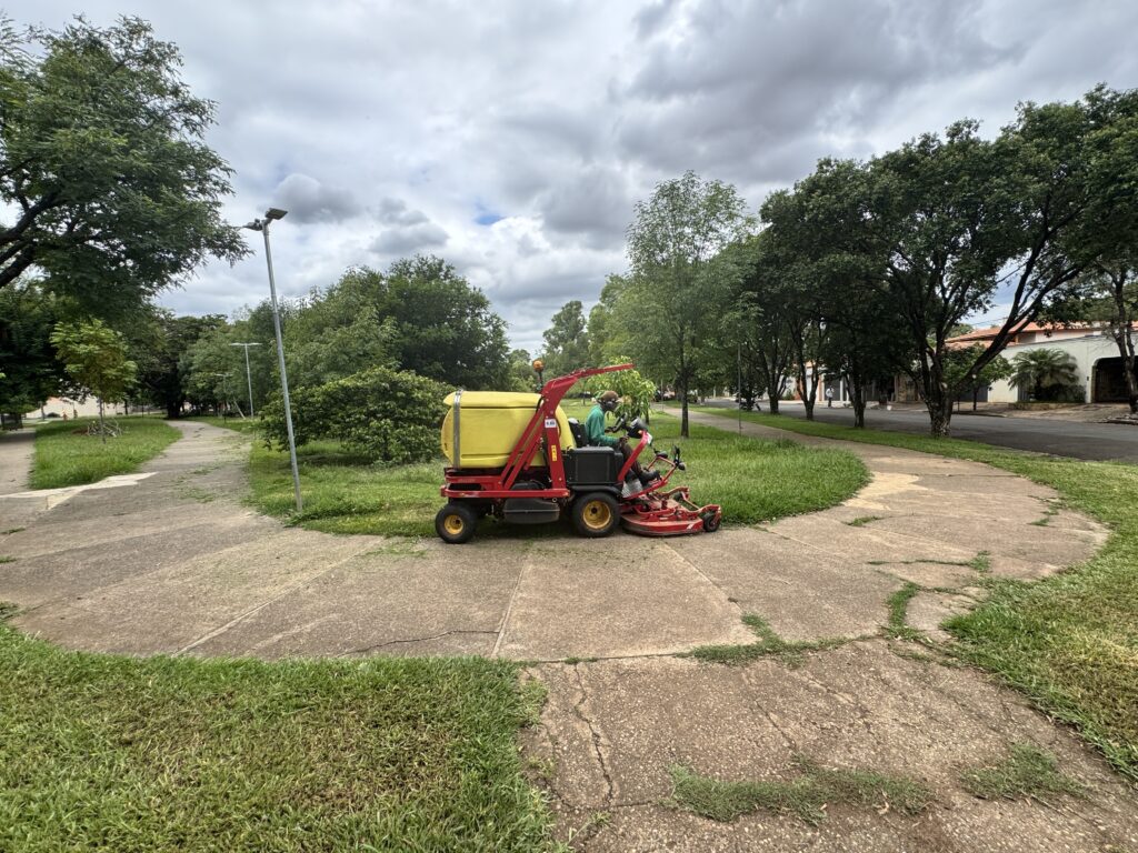 A imagem mostra um trabalhador em um cortador de grama vermelho e amarelo com um tanque amarelo, aparando a grama em uma área com caminhos de concreto. O cortador de grama está em um caminho de concreto que se divide em duas direções. Há árvores verdes e exuberantes ao redor, e o céu está nublado. A grama que está sendo cortada é verde e parece estar crescendo sobre o concreto em alguns lugares. O trabalhador está usando um capacete e fones de ouvido.