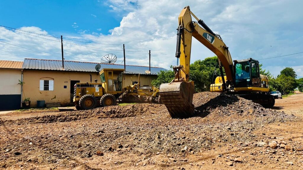 A imagem mostra um canteiro de obras com várias máquinas pesadas em ação. No centro, uma escavadeira amarela da marca CAT está a transferir terra para a caçamba de um caminhão basculante. A escavadeira está posicionada sobre um monte de terra, com seu braço estendido e a concha cheia de material. À esquerda, um motonivelador amarelo, também uma máquina de grande porte, está parado, com seu corpo robusto e rodas grandes visíveis. À direita do caminhão basculante, um caminhão branco de porte menor está estacionado, possivelmente aguardando para ser carregado ou para transportar o material. O cenário ao redor sugere uma área em desenvolvimento ou reforma, com terra solta e cascalho espalhados pelo chão. Ao fundo, observam-se algumas casas e árvores, indicando que a obra está localizada em uma área urbana ou periurbana. O céu está parcialmente nublado, com algumas áreas de azul claro.