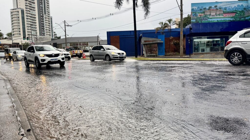 A imagem mostra uma rua urbana alagada após uma forte chuva, com grande acumulação de água no asfalto. Vários carros estão trafegando lentamente pela via inundada, incluindo um Renault Kwid branco em destaque à esquerda, com a placa OPT4A71, e um Volkswagen Polo prata logo atrás. À direita, um SUV branco aparece parcialmente na imagem. O céu está nublado, conferindo uma iluminação difusa típica de dias chuvosos, e o reflexo dos faróis dos carros pode ser visto na água da rua. Ao fundo, é possível ver prédios altos e modernos, indicando que a cena ocorre em uma grande cidade. Também há um prédio azul com um grande mural que retrata um palácio histórico e o horizonte da cidade. Um letreiro da concessionária Honda é visível em um dos edifícios. A imagem transmite a dificuldade do trânsito em função do alagamento e a situação típica de áreas urbanas em períodos de chuva intensa.