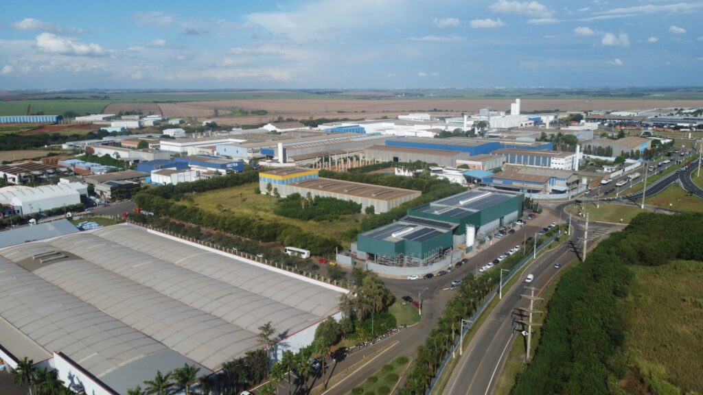 A imagem mostra uma ampla área industrial vista de cima, composta por diversos galpões e armazéns de grande porte, organizados em quadras bem definidas e interligados por ruas e estacionamentos. As construções apresentam diferentes estilos e cores de telhados, indicando a presença de várias empresas. O espaço é cercado por áreas verdes e bem planejado em termos de infraestrutura, transmitindo a ideia de um polo industrial estruturado