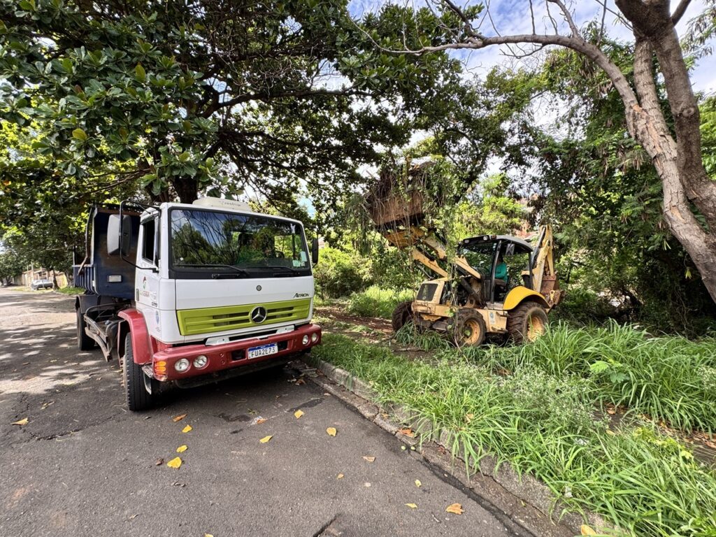 A imagem mostra um cenário de trabalho em uma área arborizada, possivelmente para limpeza ou manutenção. À esquerda, um caminhão Mercedes-Benz Atego branco com detalhes em verde e vermelho está estacionado na rua. A grade frontal do caminhão é verde, e o para-choque é vermelho. Na placa do veículo, lê-se "FUA2E73". O caminhão tem um compartimento de carga azul na parte traseira. À direita, um retroescavadeira amarela está em operação, com seu braço levantado e a caçamba cheia de galhos e folhagens, indicando que está removendo vegetação. A máquina está posicionada sobre uma área com grama alta e vegetação densa, ao lado de uma árvore grande e frondosa. O operador da máquina está visível dentro da cabine. O ambiente é ensolarado, com luz filtrada pelas árvores, e o céu azul com algumas nuvens pode ser visto entre os galhos. Há folhas caídas no asfalto, sugerindo que o trabalho pode estar relacionado à poda de árvores ou à limpeza de detritos naturais.
